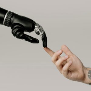 Eine menschliche Hand mit Tätowierungen berührt sanft die Fingerspitze einer schwarzen, futuristischen Roboterhand. Das Bild symbolisiert die Verbindung zwischen Mensch und Technologie.