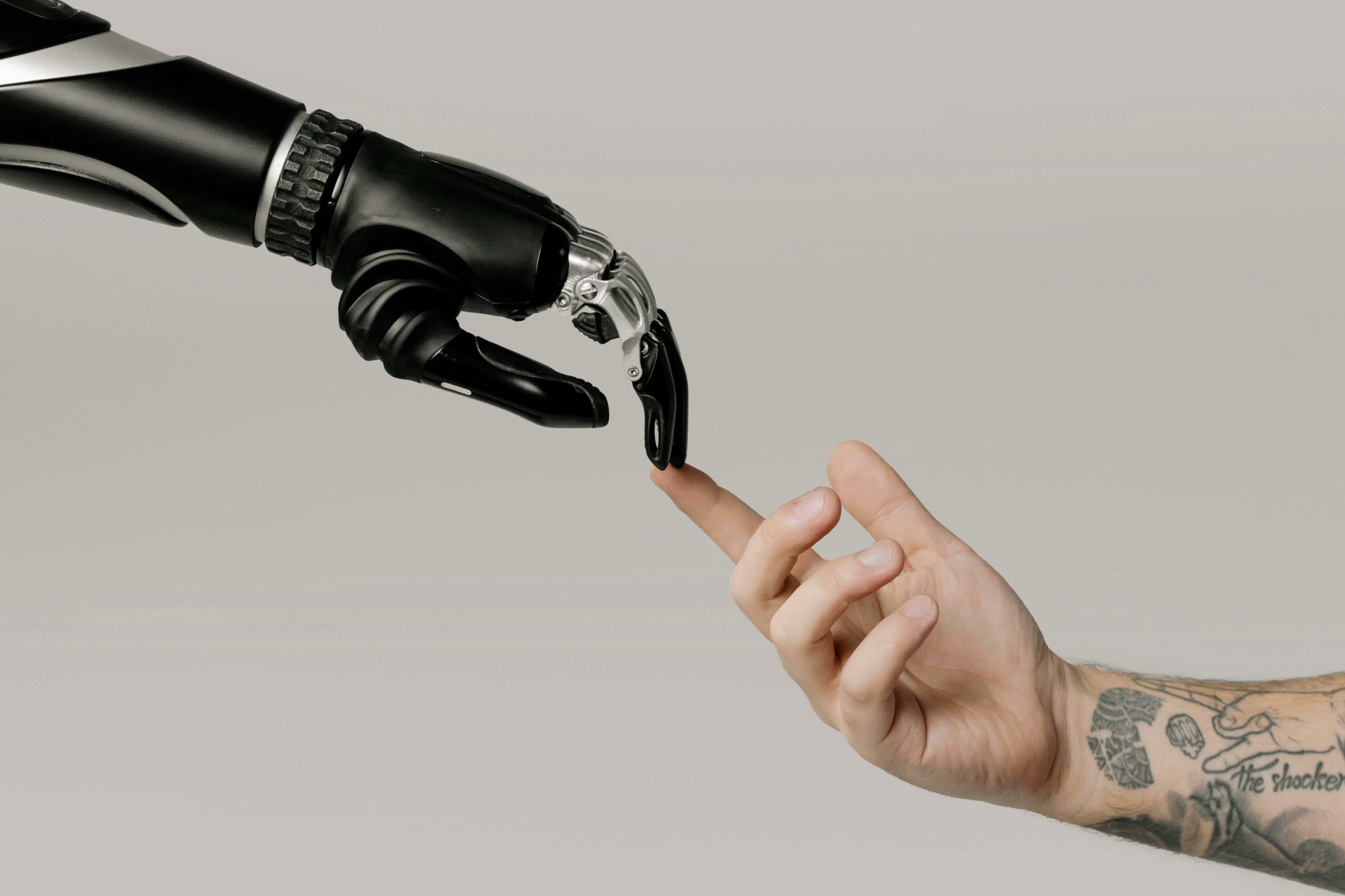 Eine menschliche Hand mit Tätowierungen berührt sanft die Fingerspitze einer schwarzen, futuristischen Roboterhand. Das Bild symbolisiert die Verbindung zwischen Mensch und Technologie.